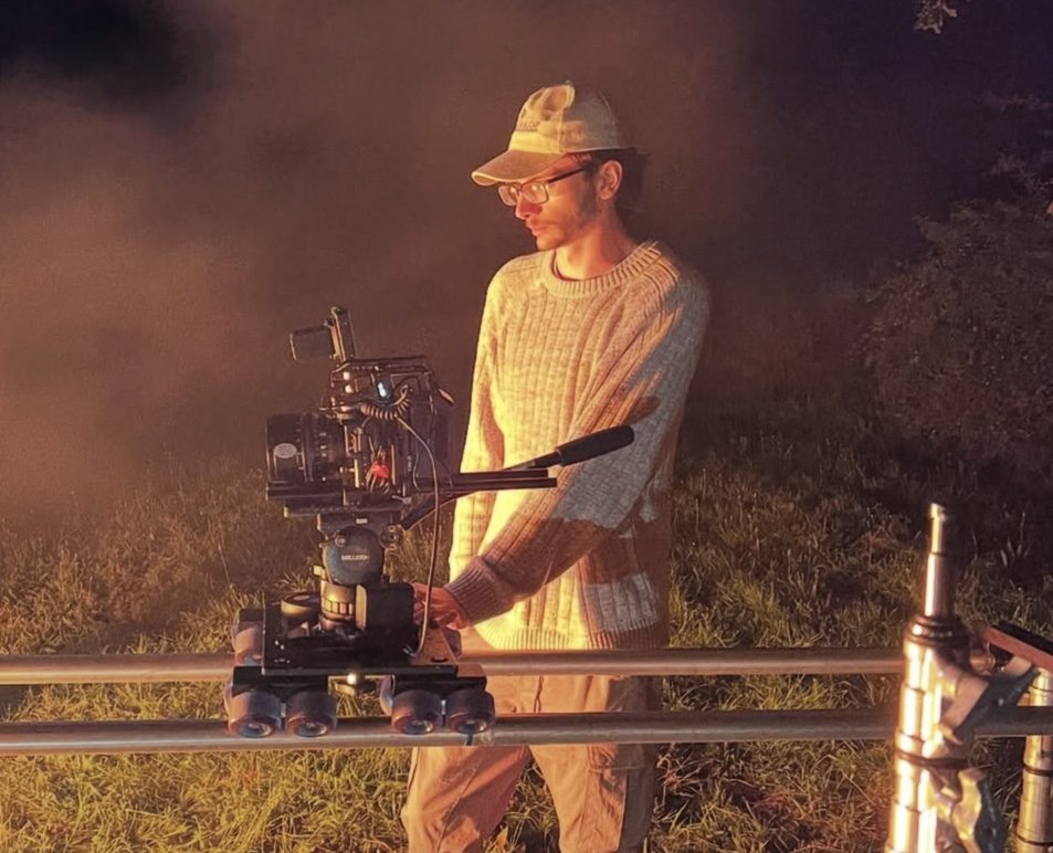 Milo Herbst operating a cinema camera on a dolly track at night, lit by warm tungsten light through haze